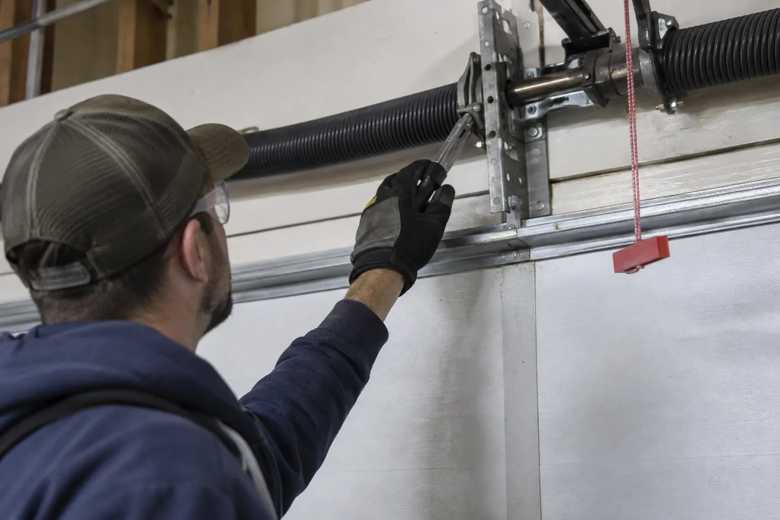 Technician in a cap reaching a gloved hand with a tool toward a coiled spring and bracket above a garage door. — High Park